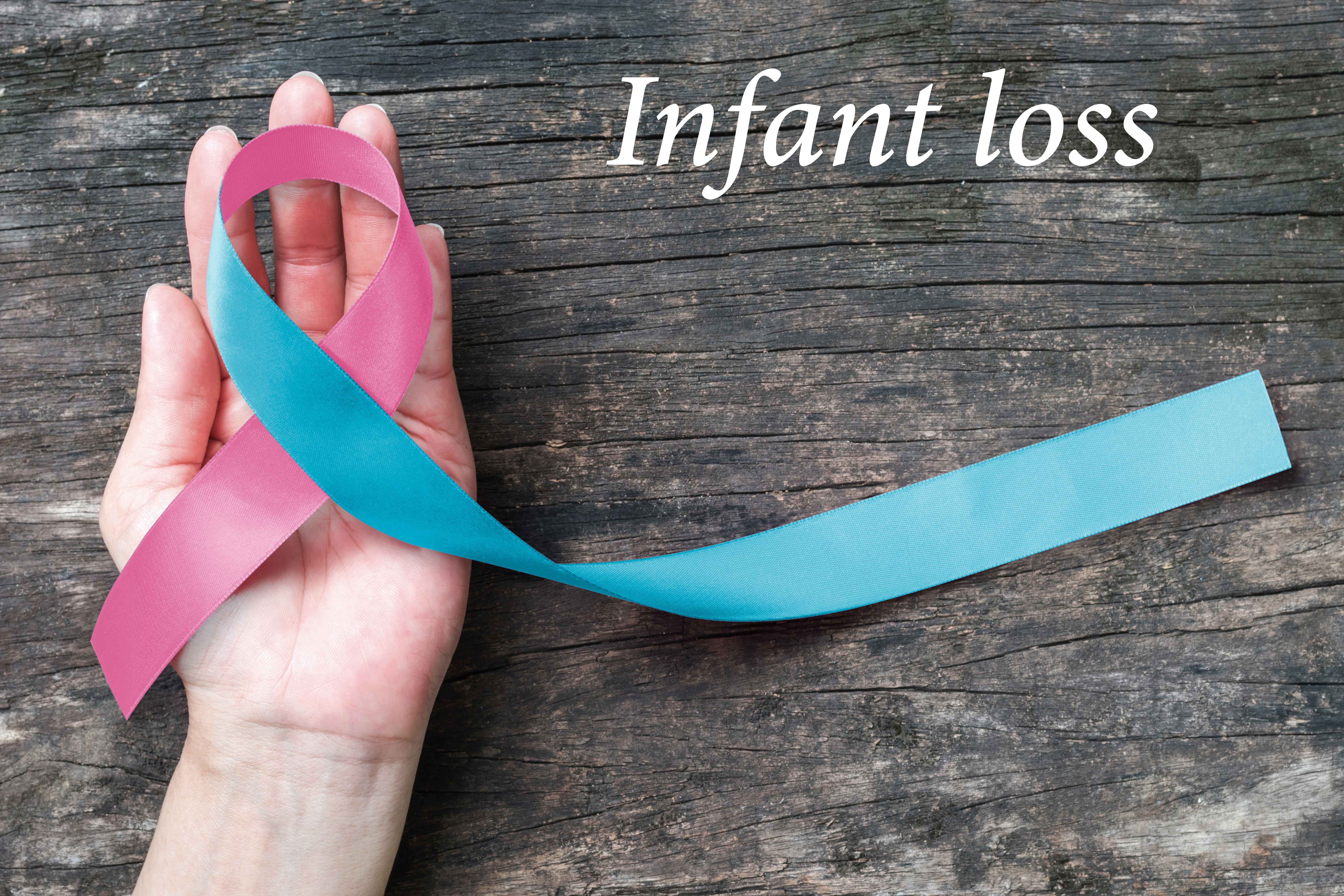 A pink and blue bonnet lays on a hand sitting on an untreated wood background. The words 'Infant loss' lay across the top right of the image.