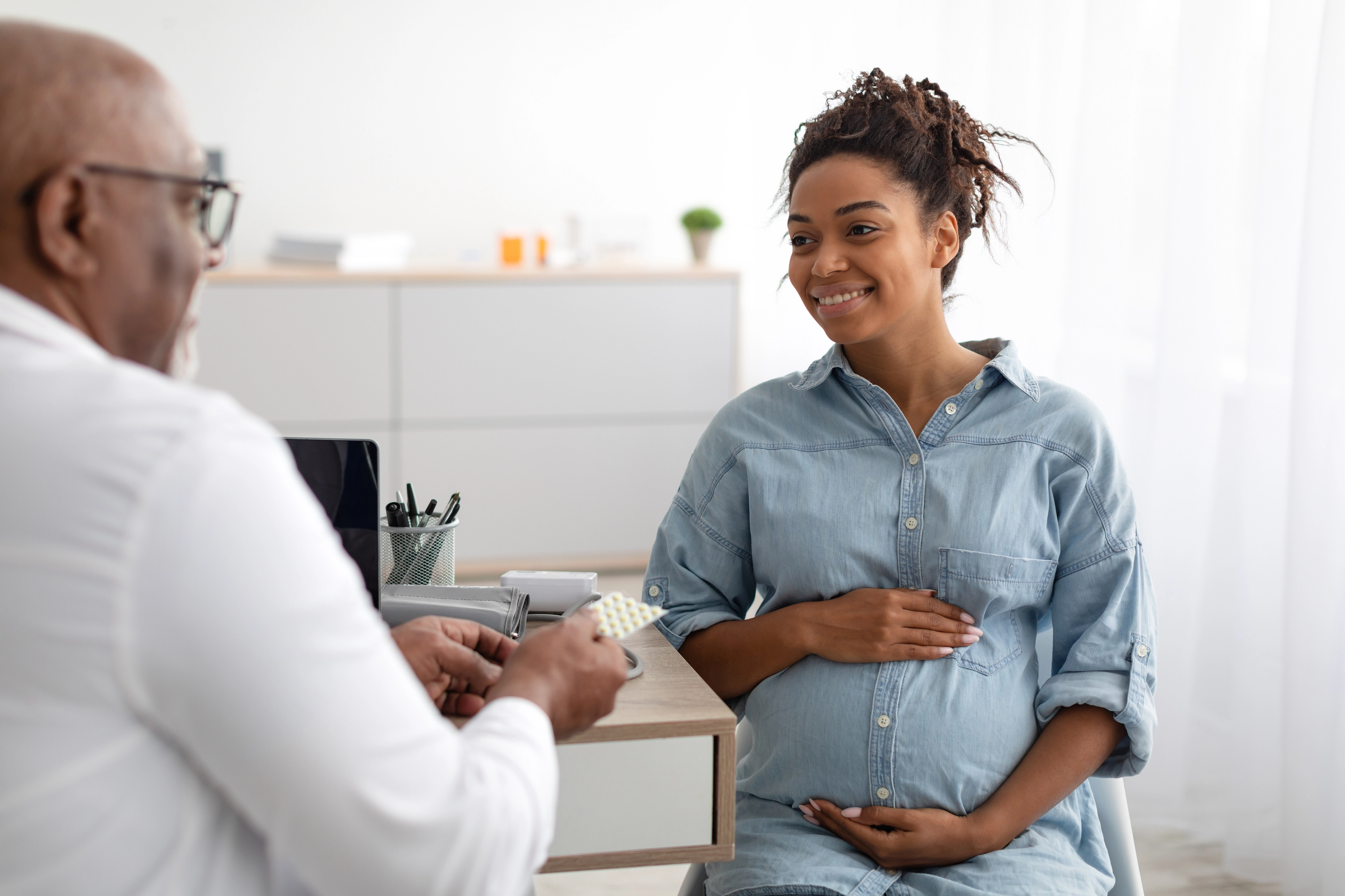 A pregnant woman with a denim shirt holds her belly while smiling at her doctor. The doctor is handing her pills and smiling.